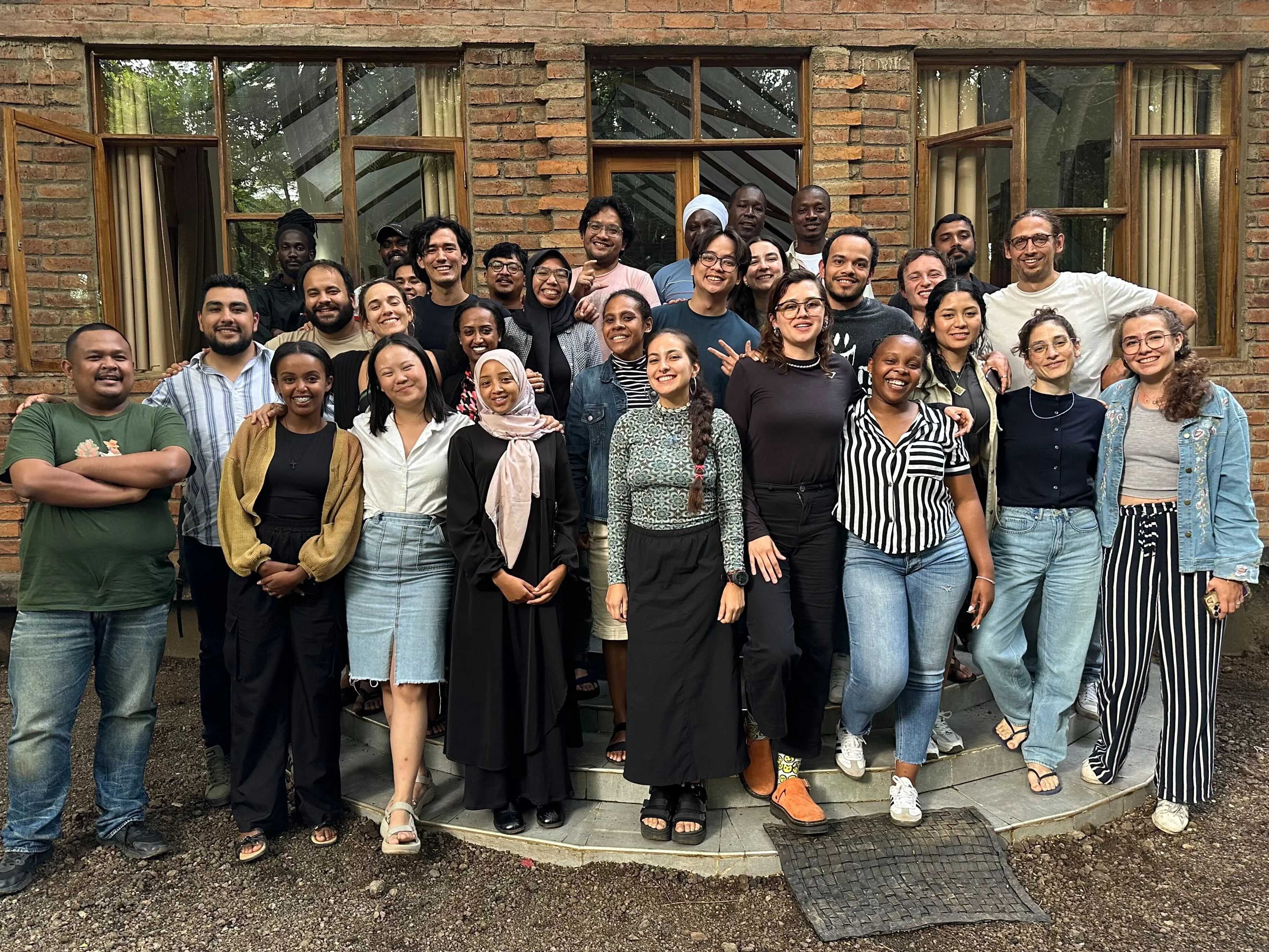 A diverse group of approximately 30 people posing together outdoors in front of a brick building with large glass windows, smiling at the camera at an ops team meeting.